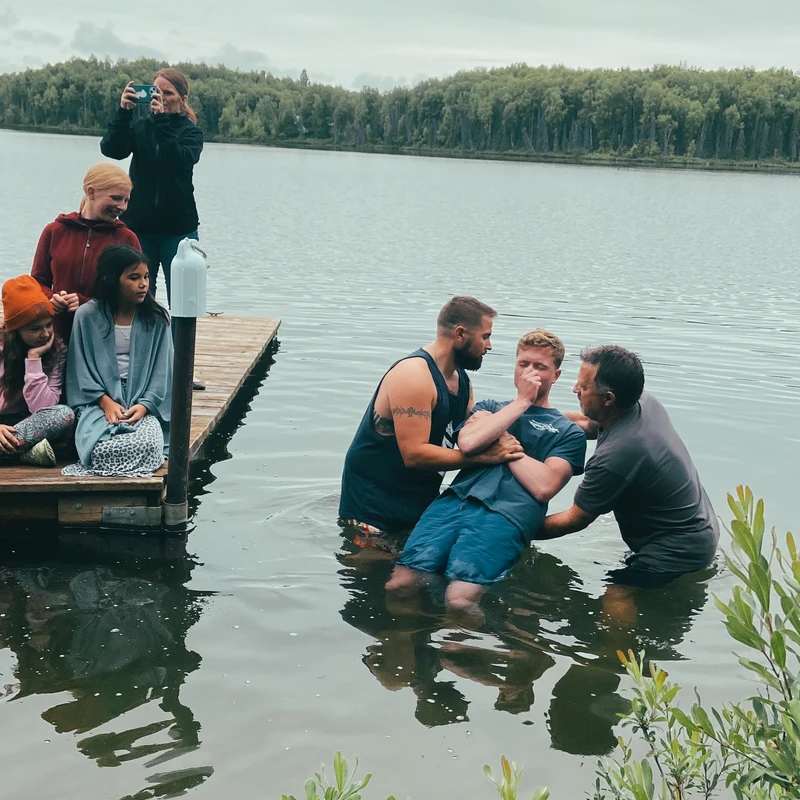 Two men baptize a young man in a lake while a group of people, including children, watch from a wooden dock. The weather appears overcast.